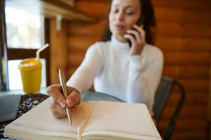 focus Aan een pen in vrouw hand- schrijven Aan een dagboek terwijl onderhandelen zittend in de buurt venster Bij een houten cafe foto