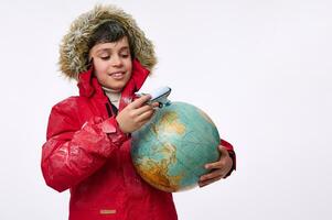 in de omgeving van de wereld reizen en winter toerisme concept. mooi kind, 10 jaren oud jongen in helder rood sneeuw gedekt naar beneden jasje spelen met een vliegtuig speelgoed- model- en wereldbol, geïsoleerd Aan wit achtergrond foto