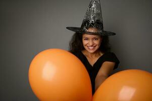 Afrikaanse Amerikaans vrouw gekleed in zwart en heks hoed gluurt door de oranje opgeblazen ballonnen en glimlacht met een mooi toothy glimlach op zoek Bij de camera. halloween concept met kopiëren ruimte foto