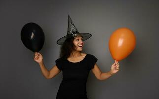 gemengd ras mooi vrouw heks in tovenaar hoed, gekleed in zwart , staat tegen grijs achtergrond met oranje en zwart lucht ballon. halloween concept met kopiëren ruimte foto