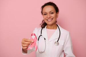prachtig Afrikaanse Amerikaans vrouw dokter met mooi toothy glimlach tonen een roze satijn lint, geïsoleerd Aan gekleurde achtergrond met kopiëren ruimte. wereld dag van strijd borst kanker, 1 st oktober concept foto