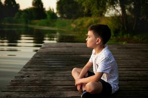 een charmant jongen resting Aan de pier en op zoek in de afstand tegen de backdrop van de rivier- bank. camping, zomer thema's. foto