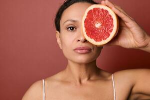 aantrekkelijk vrouw met perfect gezond huid houdt een grapefruit in voorkant van haar oog, aan het bedekken met het voor de helft van haar mooi gezicht. gebruik makend van natuurlijk biologisch fabriek gebaseerd ingrediënten voor huid zorg schoonheidsmiddelen foto