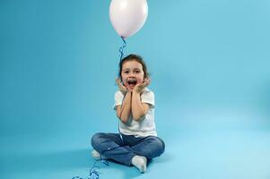 schattig weinig meisje zit Aan een blauw achtergrond en verheugt zich in een verrassing voor haar verjaardag uitdrukking van blij emoties foto