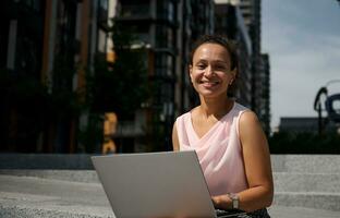 detailopname portret van een vrolijk mooi vrouw van gemengd ras werken Aan laptop zittend Aan de stappen in de stad en glimlachen op zoek Bij camera. bedrijf, ver weg afgelegen werk en opstarten concept foto