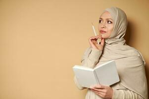 jong mooi moslim vrouw in hijab op zoek naar de kant met een nadenkend kijken Holding een wit kladblok en een potlood Bij haar kin. foto