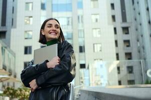 geslaagd vrolijk mooi vrouw, bedrijf persoon, freelancer, ondernemer, tekstschrijver Holding laptop in haar handen en schattig glimlacht op zoek terzijde Aan modern kantoor zakelijke gebouwen achtergrond foto