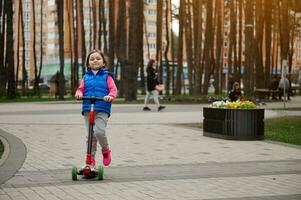 vol lengte portret van een Europese schattig kind meisje rijden een trap scooter in de park Aan een zonnig dag in vroeg herfst of de lente. gelukkig kinderjaren concept foto