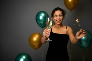 vrolijk vrouw in zwart avond jurk houdt een glas van Champagne en sterretjes, poses tegen grijs achtergrond met mooi decoratie van goud en groen lucht ballonnen. kerstmis, verjaardag concept foto