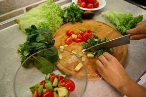 vrouw chef hakken rauw groenten en rucola bladeren Aan een houten keuken bord, voorbereidingen treffen rauw veganistisch salade Bij huis keuken foto