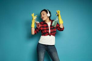 jong Afrikaanse vrouw vervelend rood plaid overhemd en blauw jeans met hoofdtelefoons Aan haar hoofd zingt, poseren Aan een blauw achtergrond met schoonmaak sponzen in haar handen foto