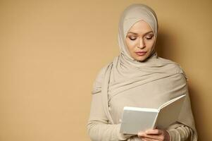 portret van een stil moslim vrouw vervelend een hijab en leunend haar hoofd naar beneden lezing een wit boek in haar handen foto