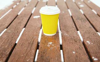 geel meenemen kop van heet drankje, koffie of thee Aan houten bank in een besneeuwd winter park foto