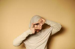 een moslim vrouw in hijab looks Bij de camera door haar vingers framing haar gezicht staand Aan beige achtergrond met kopiëren ruimte foto