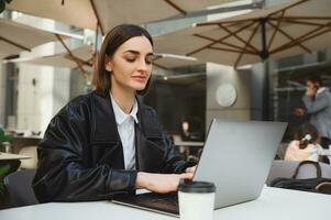 zelfverzekerd jong bedrijf vrouw, tekstschrijver, ontwikkelaar, freelancer, journalist werken van een afstand Aan laptop, schrijven tekst, genieten van ver weg werk in buiten zomer cafetaria Aan mooi vroeg voorjaar dag foto