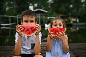 glimlachen schattig kinderen aan het bedekken mond met gesneden watermeloen, zittend Aan de pier, genieten van platteland zomer vakanties Bij avond. gelukkig kinderachtig emoties Aan de gezichten van aanbiddelijk jongen en meisje foto