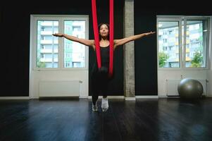 mooi Afrikaanse sportief vrouw hangende Aan een rood hangmat met uitgestrekt handen in een geschiktheid en yoga studio. slank lichaam jong vrouw beoefenen antenne vliegend yoga met anti zwaartekracht foto