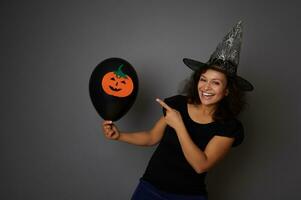 mooi vrouw gekleed in halloween carnaval heks kleren, points Aan zwart ballon met een vilt gesneden pompoen, glimlacht toothy glimlach op zoek Bij camera, geïsoleerd over- grijs achtergrond met kopiëren ruimte foto