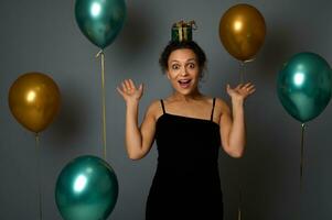 vrolijk vrouw in avond zwart kleding verheugt zich op zoek Bij camera met een geschenk Aan haar hoofd, poses tegen grijs muur achtergrond met schitteren gouden groen lucht ballonnen. kerstmis, verjaardag concept foto