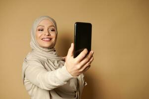 elegant Arabisch vrouw met mooi toothy glimlach en gedekt hoofd maken selfie staand tegen een beige achtergrond met kopiëren ruimte. foto