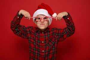 knap schattig kind jongen in de kerstman claus hoed en plaid overhemd looks Bij de camera door Kerstmis zoet snoep wandelstokken, Holding hen imiteren bril, poseren Aan rood achtergrond met kopiëren ruimte voor advertentie foto