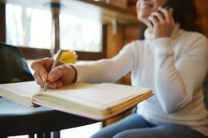 detailopname van vrouw hand- Holding een zilver pen en schrijven Aan een zuivel Bij een houten cafetaria bar foto