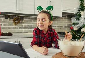 gelukkig positief weinig meisje met elf hoepel Aan haar hoofd, gekleed in rood en groen plaid jurk pratend door video telefoontje en opname video, staand door tafel en menging ingrediënten voor Kerstmis gebakje foto