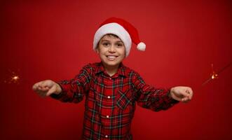 knap aanbiddelijk preteen jongen, mooi kind in de kerstman claus hoed en geruit overhemd geniet de Kerstmis partij, Toneelstukken met Bengalen lichten, sterretjes, geïsoleerd O rood achtergrond met kopiëren ruimte voor advertentie foto