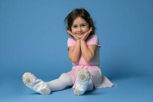 portret van een aanbiddelijk kind meisje ballerina schattig glimlachen terwijl zittend Aan blauw achtergrond foto