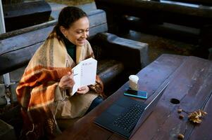 mooi vrouw glimlacht tonen met pen Aan blanco blanco bladzijde met Te doen lijst gedurende video conferentie Aan laptop, houdt warm door omhulsel haarzelf in wollen geruit deken, zittend buitenshuis Aan houten bank foto