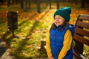 vrolijk aanbiddelijk verheugd weinig meisje in warm knus kleurrijk kleren zittend Aan houten park bank met Gesloten ogen, genieten van herfst het weer. mooi zon stralen vallend door bomen. gelukkig kinderjaren foto