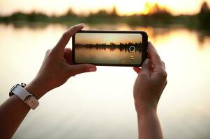 detailopname van handen nemen een fotografie van een mooi zonsondergang Aan de oever van het meer achtergrond. zomer thema's. mobiel telefoon in leven visie foto