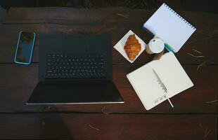 vlak leggen samenstelling van bedrijf en kantoor artikelen. laptop, smartphone, Open schrijfboek, kladblok en vers gebakken croissant met meenemen papier mok van heet koffie Aan een houten tafel oppervlak. hoog hoek visie foto