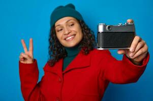 focus Aan retro stijl fotografisch uitrusting wijnoogst camera in de handen van wazig mooi vrouw, tonen vrede teken en glimlachen met toothy glimlach terwijl nemen selfie, tegen blauw gekleurde achtergrond foto