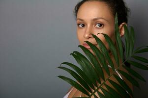 mooi spaans vrouw met natuurlijk bedenken en schoon vers gezond huid covers haar gezicht met palm blad, op zoek vol vertrouwen Bij camera, poseren tegen grijs muur achtergrond, kopiëren ruimte. spa concept foto