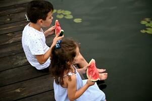 schattig kinderen met plakjes van watermeloen in hun handen, zittend Aan de pier met hun poten verlaagd in de water en genieten van zomer avonden in de platteland. foto