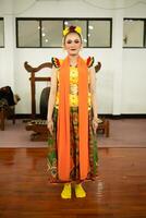 een traditioneel Indonesisch danser staand in een geel kostuum en een oranje sjaal hangende naar beneden haar lichaam foto