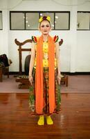 een traditioneel Indonesisch danser staand in een geel kostuum en een oranje sjaal hangende naar beneden haar lichaam foto