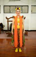 een traditioneel Indonesisch danser staand in een geel kostuum en een oranje sjaal hangende naar beneden haar lichaam foto