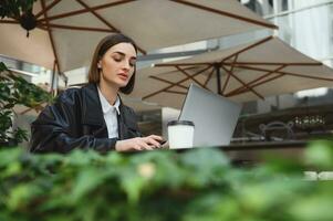 mooi geconcentreerd Europese brunette, mooi vrouw, tekstschrijver, freelancer werken Aan laptop locatie in zomer terras van een cafetaria. ver weg afgelegen online bedrijf concept foto