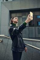 modieus jong zelfverzekerd vrouw met elegant kort kapsel vervelend zwart eco leer jasje poseren tegen grijs steen gebouw met een smartphone in haar uitgestrekt hand, nemen selfie foto