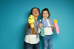 mooi jong vrouw, huisvrouw regisseert schoonmaak sprays net zo als het schieten van een pistool, staand De volgende naar haar weinig aanbiddelijk dochter . huis werk en klusjes concept foto