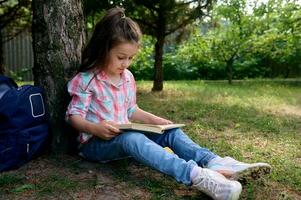 weinig school- meisje leest een boek in de park, zittend onder boom met haar rugzak. kinderen. eruditie. onderwijs. levensstijl foto