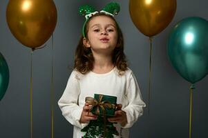 mooi kind meisje in elf hoepel Aan haar hoofd glimlacht op zoek Bij camera, poses tegen luxe glimmend lucht ballonnen Holding een Kerstmis Cadeau verpakt in glimmend groen omhulsel geschenk papier met gouden boog foto
