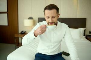 geslaagd welvarend midden- oud Kaukasisch zakenman in bedrijf gewoontjes kleding drinken koffie in hotel appartement gedurende bedrijf reis. vers gebrouwen koffie voor de perfect begin naar de dag foto