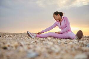jong vrouw zittend Aan de strand gefocust Aan uitrekken haar lichaam gedurende ochtend- training Bij zonsopkomst foto