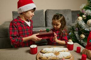 aanbiddelijk kinderen, jongen in de kerstman hoed en meisje zittend Bij een tafel met vers gebakken traditioneel Duitse stollen brood en een heet chocola drinken met marshmallows, en geven elk andere cadeaus voor kerstmis. foto