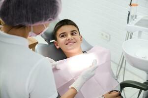 vrolijk donker haar schooljongen aan het liegen Aan tandarts stoel, glimlacht met mooi toothy glimlach op zoek Bij tandheelkundig assistent voorbereidingen treffen hem voor regelmatig medisch controle. tandheelkunde, mondeling zorg hygiëne concept foto