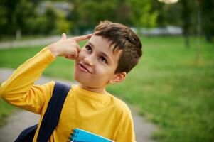 een charmant blij vrolijk schooljongen houdt zijn hand- naar zijn tempel imiteren een pistool, net zo een teken van vermoeidheid van aan het studeren na een moeilijk dag Bij school, op zoek omhoog in de stad park achtergrond foto