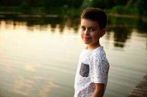 portret van een charmant jongen schattig lachend, op zoek Bij de camera Aan de achtergrond van de meer. zonsondergang en een lijn van bomen zijn weerspiegeld in de meer water foto